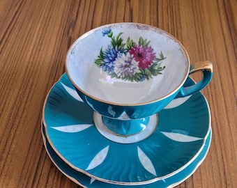 Puede incluir: Un juego de taza y platillo de té de color turquesa con un diseño floral. La taza tiene un interior blanco con un borde dorado y un diseño floral en el centro. El platillo es turquesa con un borde blanco y un ribete dorado.