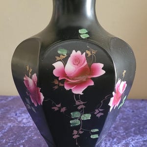Puede incluir: Jarrón negro con forma hexagonal y parte superior acampanada. El jarrón está decorado con rosas rosas y hojas verdes. El diseño floral destaca sobre el fondo negro. El jarrón probablemente esté hecho de cerámica o porcelana.