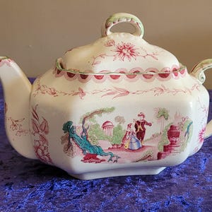 May include: Cream-colored ceramic teapot with a lid, handle, and spout. The teapot is decorated with pink floral patterns and a pastoral scene with figures in a garden. The rim and handle have green accents.