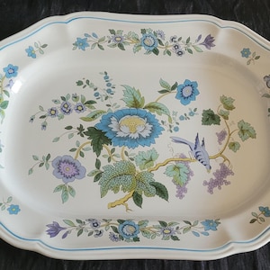 Könnte beinhalten: Eine ovale Servierplatte aus Keramik mit gewelltem Rand und einer dünnen blauen Umrandung. In der Mitte befindet sich eine detaillierte Abbildung einer großen blauen Blume, grünen Blättern, violetten Trauben und einem kleinen blauen Vogel.