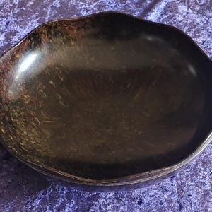 Puede incluir: Un cuenco marrón oscuro de forma irregular con un acabado brillante. El borde tiene un diseño ondulado. El interior del cuenco es de color marrón oscuro liso, mientras que el borde exterior tiene un patrón moteado de marrón, rojo y amarillo.