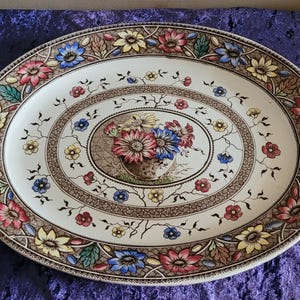 May include: Oval serving platter with a floral design. The off-white ceramic platter features a brown border with colourful flowers in red, blue, and yellow. The centre has a floral bouquet in a vase. A decorative piece for serving food.