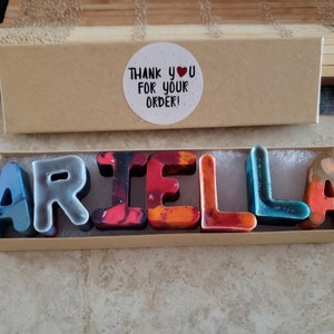 Puede incluir: Una caja de cartón con una pegatina blanca que dice "¡Gracias por tu pedido!" y un corazón. La caja contiene un juego de letras de crayones de colores que deletrean el nombre "ARIEL LA".