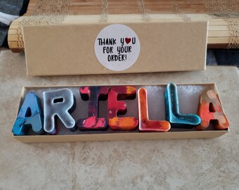 Puede incluir: Una caja de cartón con una pegatina blanca que dice "¡Gracias por tu pedido!" y un corazón. La caja contiene un juego de letras de crayones de colores que deletrean el nombre "ARIEL LA".
