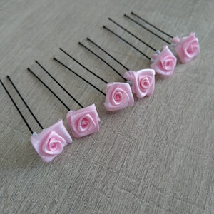 Op de afbeelding: Een verzameling roze stoffen rozen haarspelden. Elke speld heeft een kleine, vierkante roos van zachte, lichtroze stof, bevestigd aan een zwarte metalen speld. De rozen zijn in een rij gerangschikt en tonen hun delicate ontwerp.