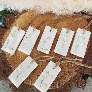 Pode incluir: Conjunto de seis derretes de cera retangulares brancos com delicados desenhos florais. Cada derrete é amarrado com um barbante de juta natural e apresenta um padrão floral diferente.