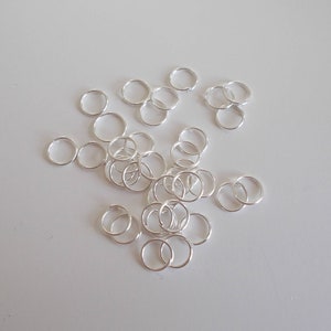 Op de afbeelding: Een verzameling kleine, glanzende, zilveren verbindingsringen verspreid over een witte ondergrond. De ringen zijn cirkelvormig en lijken van metaal te zijn, mogelijk sterling zilver. Ze worden waarschijnlijk gebruikt voor het maken van sieraden of knutselen.