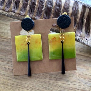 Peut inclure: Une paire de boucles d'oreilles composée d'un clou circulaire noir texturé relié à un pendentif carré avec un dégradé de jaune et de vert. Une fine breloque noire allongée est suspendue au centre de chaque carré par des anneaux dorés.
