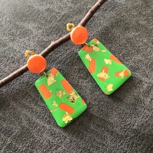 Può includere: Un paio di orecchini pendenti con un perno circolare arancione brillante attaccato a un pendente rettangolare verde. I pendenti rettangolari sono decorati con accenti rettangolari arancioni e scaglie di foglia d'oro sparse sulla superficie.