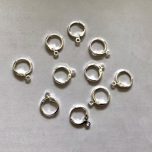 Pode incluir: Uma coleção de nove bases para brincos de argola em metal prateado dispostas de forma dispersa. Cada componente circular apresenta um mecanismo de fecho de alavanca e um pequeno elo na parte inferior para prender pingentes ou berloques.