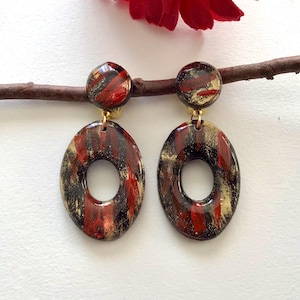 May include: A pair of drop earrings featuring a circular stud top and a larger oval-shaped pendant. The surface of each earring has a glossy finish with a marbled pattern in shades of deep black, vibrant red, and metallic gold throughout the pieces.
