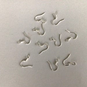 Peut inclure: Un ensemble de neuf crochets de boucles d'oreilles en métal argenté, chacun présentant un design en fil courbé avec une petite boucle circulaire à la base pour attacher des éléments de bijoux. Ces apprêts sont disposés de manière éparse.