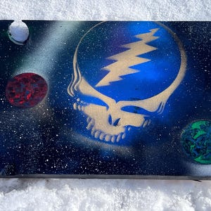 May include: A black wooden canvas with a blue and gold Grateful Dead Steal Your Face stencil design. The background is a dark blue with white speckles, and there are two small red and green circles in the corners.
