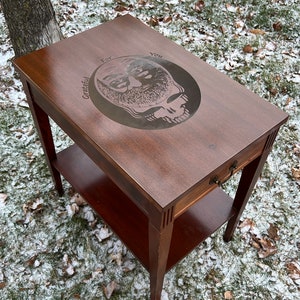 May include: A dark brown wooden end table with a single drawer and a lower shelf. The top of the table is engraved with a circular design featuring a skull and the words "For Grateful You".
