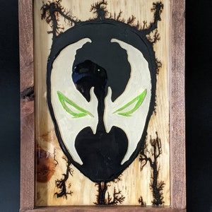 May include: A black and white resin art piece of a superhero mask with green eyes. The mask is set in a wooden frame with a wood-burned background.