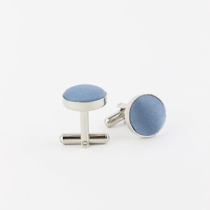May include: A pair of silver cufflinks with a round, light blue fabric center.
