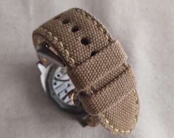 May include: A tan canvas watch strap with yellow stitching. The strap is folded over and shows the stitching detail.