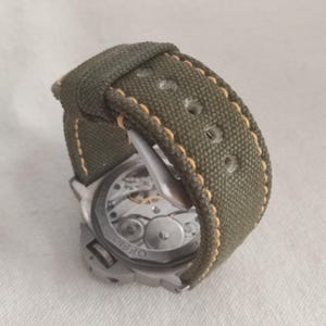 May include: A silver watch with a green canvas strap and a visible mechanical movement. The strap has a yellow stitching detail.