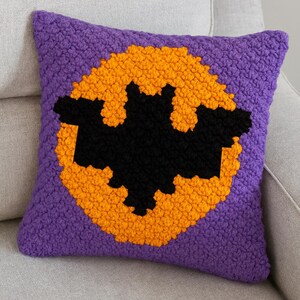 May include: A square cushion with a Halloween theme. The cushion features a black bat design on an orange circle, set against a purple background. The cushion is made of a textured fabric.