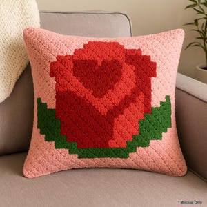 May include: A square, pink crochet cushion with a large red rose design and green leaves. The rose is centrally placed and made of textured yarn, creating a three-dimensional effect. The cushion is on a grey sofa.