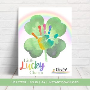 Pode incluir: Uma impressão em aquarela com um trevo verde de quatro folhas com impressões de mãos coloridas no centro e um arco-íris arqueado acima. O texto "Little Lucky Charm" está escrito em cores do arco-íris, com o nome "Oliver" abaixo.