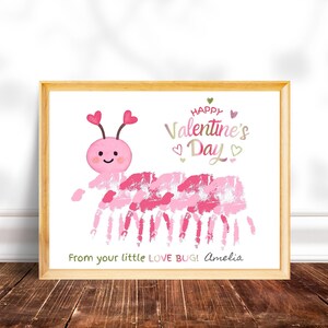 Op de afbeelding: Een ingelijst Valentijnsdagkunstwerk met een roze handafdrukrups met een lachend gezicht en hartantennes. De tekst luidt "Happy Valentine's Day" en "From your little LOVE BUG!" met een naam eronder.