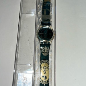 Puede incluir: Un reloj Swatch vintage con una correa verde y dorada, con gráficos temáticos de los Juegos Olímpicos. La esfera del reloj es oscura con detalles dorados. La correa muestra el texto "London 1948" y "Melbourne 1956".