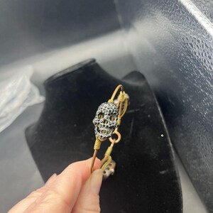 May include: A gold-toned wire bangle bracelet featuring a central skull charm encrusted with small black crystals. The wire is wrapped around the skull charm, and the bracelet includes additional metallic accents along the circular band structure.