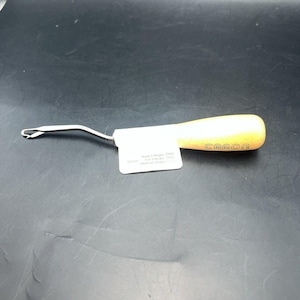 May include: A latch hook tool with a light-colored wooden handle featuring the brand name CARON printed in black. The metal hook has a curved neck and a hinged latch mechanism. A white paper tag is attached to the handle near the metal shaft.