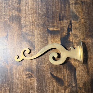 May include: A brass wall-mounted hook featuring a curved, scroll-like design with a flat circular base plate at one end and a small hook at the other. The metal has a smooth, matte finish with a warm, golden-yellow metallic tone.