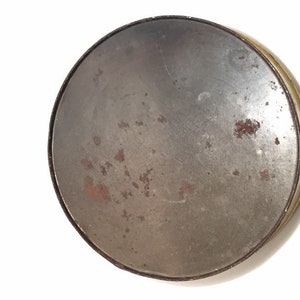 Puede incluir: Una lata redonda de metal con la parte superior ligeramente abollada. La lata es de color dorado opaco y descolorido con algunas manchas de &oacute;xido.