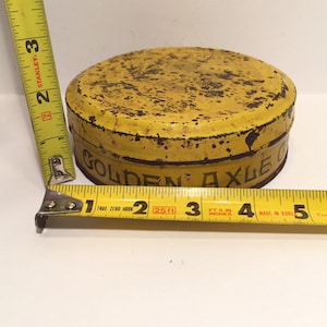 Puede incluir: Una lata de metal amarilla con las palabras "Golden Axle Grease" impresas en el lateral. La lata tiene un di&aacute;metro de aproximadamente 7,5 cm y presenta signos de &oacute;xido.