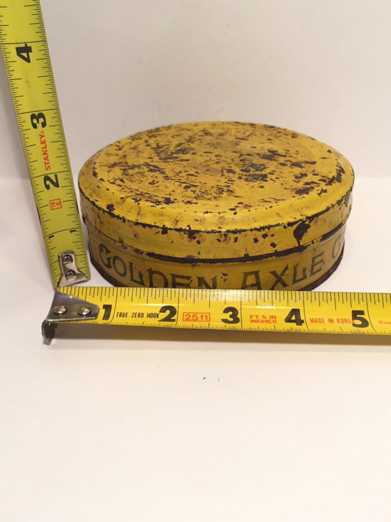 Puede incluir: Una lata de metal amarilla con las palabras "Golden Axle Grease" impresas en el lateral. La lata tiene un di&aacute;metro de aproximadamente 7,5 cm y presenta signos de &oacute;xido.