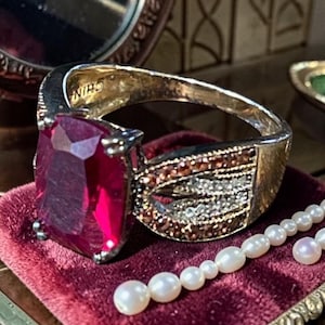 May include: A gold-tone ring featuring a large, rectangular, faceted deep pink gemstone. The band is embellished with rows of small, sparkling brown and clear stones in a decorative pattern. The interior of the band is stamped with the word CHINA.