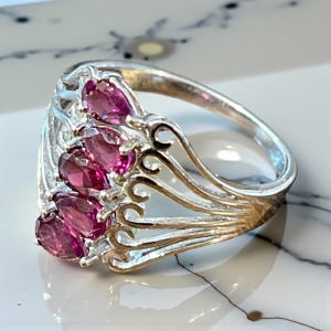 May include: A silver ring featuring a cluster of oval-cut, deep pink gemstones. The ring has an ornate, openwork design with delicate curves and a polished finish. The gemstones are set in a row, creating a striking visual effect.