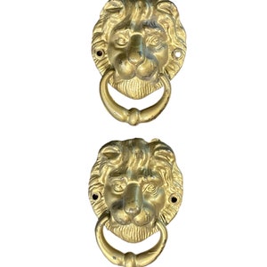 May include: Two gold lion head door knockers with a ring handle. The lion heads are detailed with a mane and whiskers.
