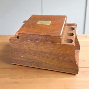 May include: A vintage wooden tobacco box with a hinged lid and three circular holes. The box is a warm brown color with a rectangular lid and a small, rectangular brass plate. The box is sitting on a wooden surface.