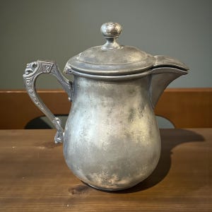 May include: Antique silver-colored pitcher with a lid and a decorative handle. The pitcher has a rounded body and a spout for pouring. The handle features ornate detailing. The lid is topped with a small, round knob. The pitcher is likely made of metal.