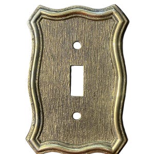 May include: Ornate, antique-style light switch cover in a gold tone. The cover has a textured surface and a decorative, curved border. It features a single toggle switch opening and two screw holes.