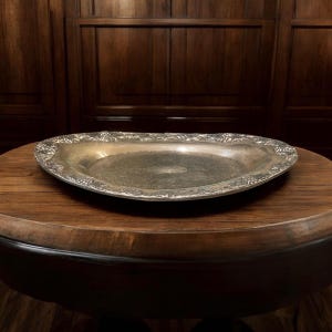 May include: Oval silver-toned serving tray with ornate floral detailing around the rim. The tray is placed on a dark wooden table, showcasing its reflective surface. A classic piece for serving or display.