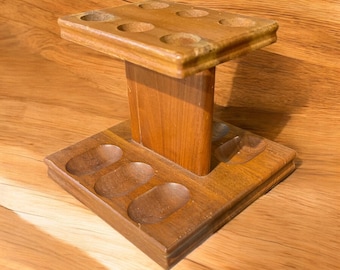 May include: A wooden pipe stand with a rectangular base and top, connected by a central support. The stand has six circular holes on top and several oval-shaped cutouts on the base, all designed to hold pipes. The wood has a warm, natural finish.