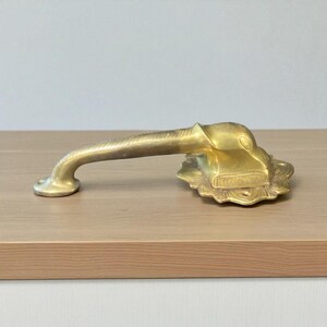 May include: A brass door handle with an ornate design. The handle features a curved bar and a decorative base with a detailed animal head. The metal has a warm, golden hue, suggesting an antique or vintage style.