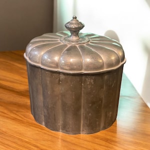 May include: A vintage metal container with a lid. The container is cylindrical with a fluted design and a decorative finial on the lid. The metal has a tarnished, aged appearance, suggesting it is an antique or vintage piece. The container sits on a wooden surface.