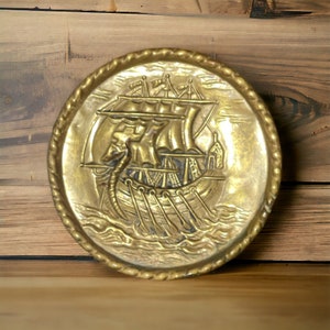 May include: A gold-toned metal wall plaque with a raised design of a ship with sails and oars. The plaque has a rope-like border.