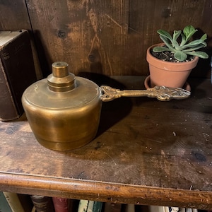May include: A vintage brass oil lamp with a decorative handle. The lamp has a round base and a cylindrical top. The handle is ornate and extends horizontally. The lamp is displayed on a wooden shelf, with a small potted plant in the background.