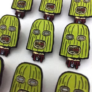 May include: A set of eight green enamel pins featuring a cartoon character wearing a green ski mask with a bloody neck. The character has gold teeth and white eyes.