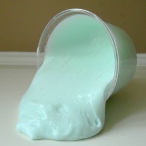 May include: A light green slime pours out of a clear plastic container onto a white surface.