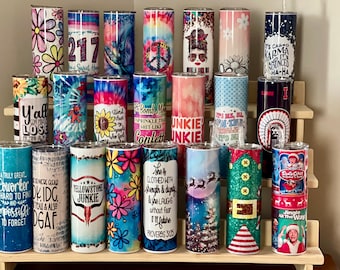 May include: A collection of various 20 oz stainless steel tumblers with different designs. The tumblers feature floral patterns, tie-dye, inspirational quotes, and holiday themes. Some have text like "217", "Yellowstone Junkie", and "Santa Claus".