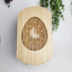 May include: A light-colored wooden box with a carved design. The design features an egg shape with a crescent moon and leaf pattern in the center, surrounded by a vine-like border. The box has a curved top and a light wood grain.