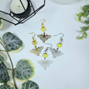 May include: Four pairs of bee-themed earrings. Two pairs are gold-toned, and two are silver-toned. Each earring features a cutout bee pendant and a yellow bead. The earrings are displayed on a white surface with greenery.